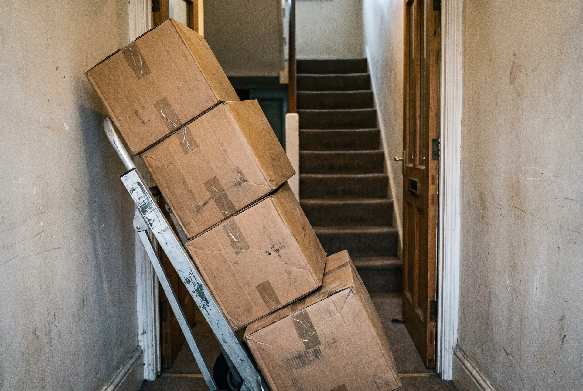 A hand truck stacked with plain cardboard boxes in a narrow hallway